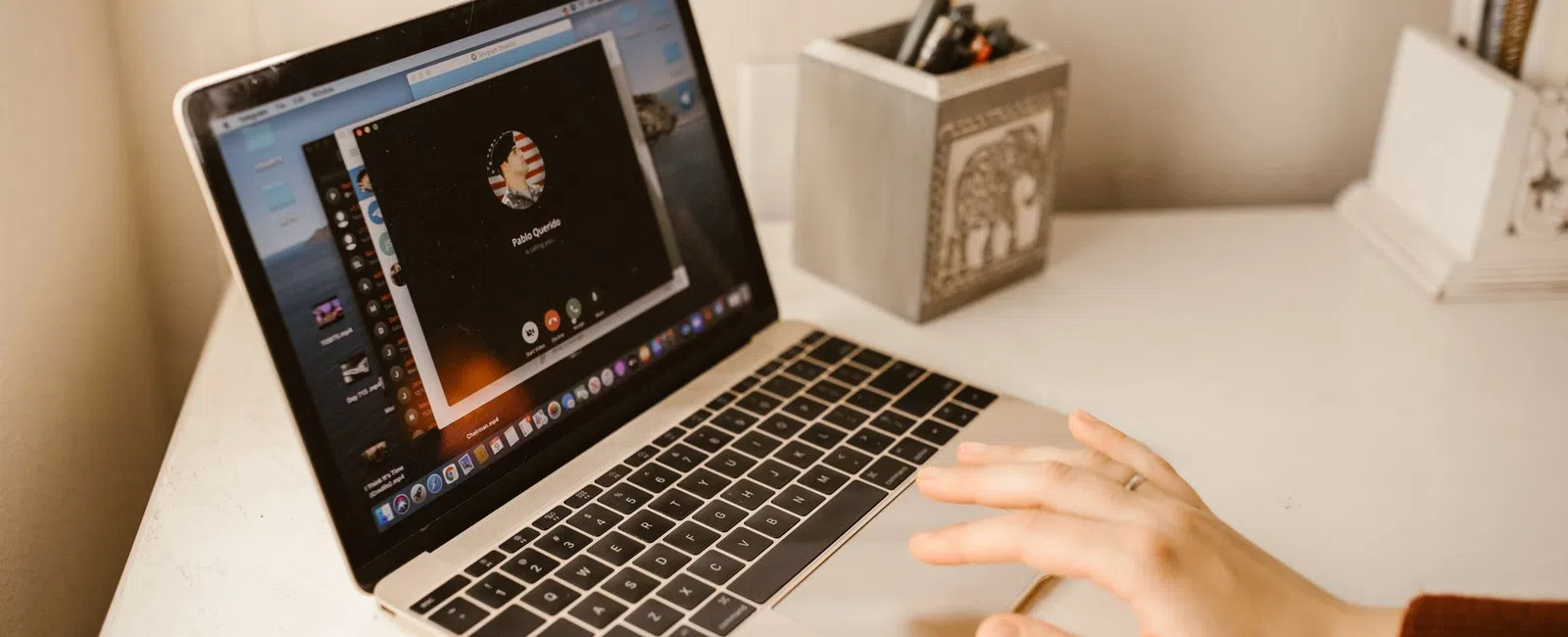 Person's hand using a MacBook with a video call screen open on a clean white desk near a pencil holder.