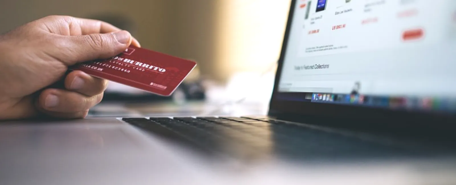 Hand holding a red credit card near a laptop keyboard with an online shopping website on screen