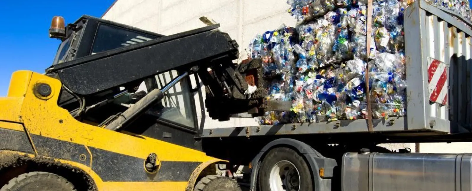 Yellow forklift loading compacted plastic bottles into a truck for recycling under a clear blue sky