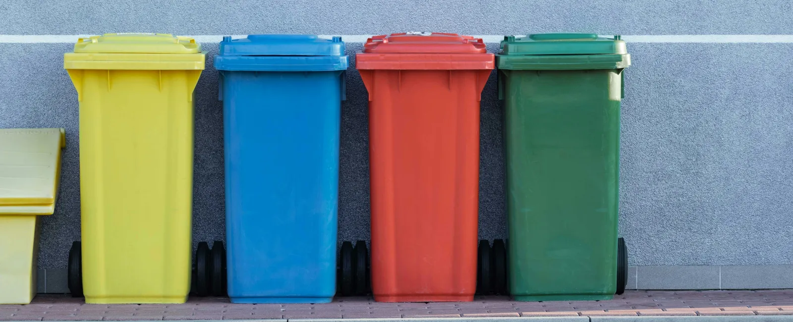 Four colorful garbage bins in yellow, blue, red, and green lined up against a gray wall on a paved street.