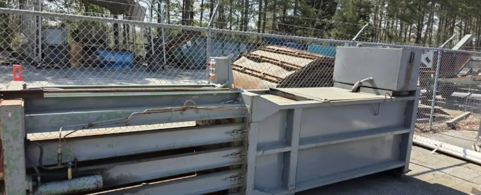 Large industrial metal baler machine outdoors near a chain-link fence and trees in daylight.