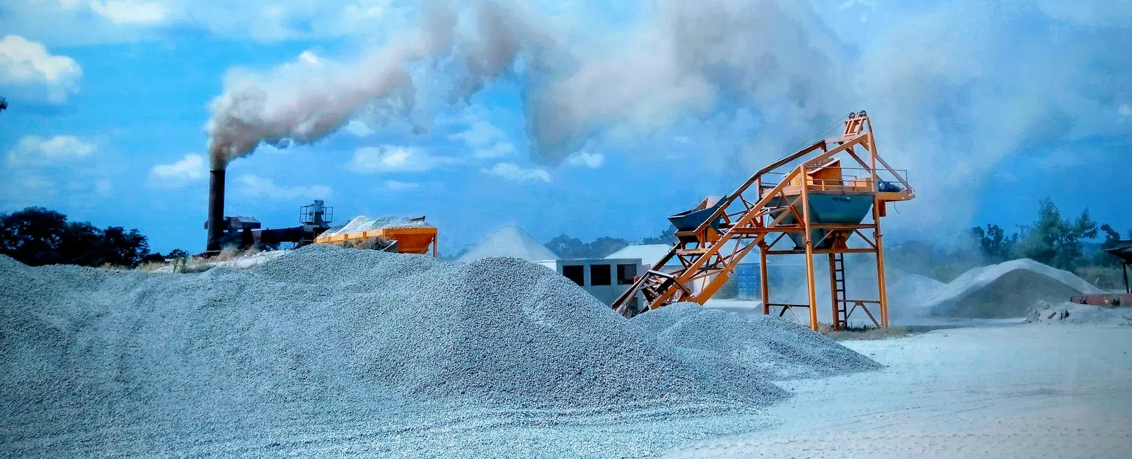 Construction site with gravel piles, conveyor belt, and factory smoke emitting under blue sky with clouds.