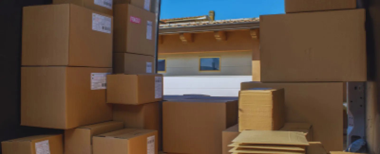 Stacked cardboard boxes inside delivery van with open door showing clear blue sky and building outside