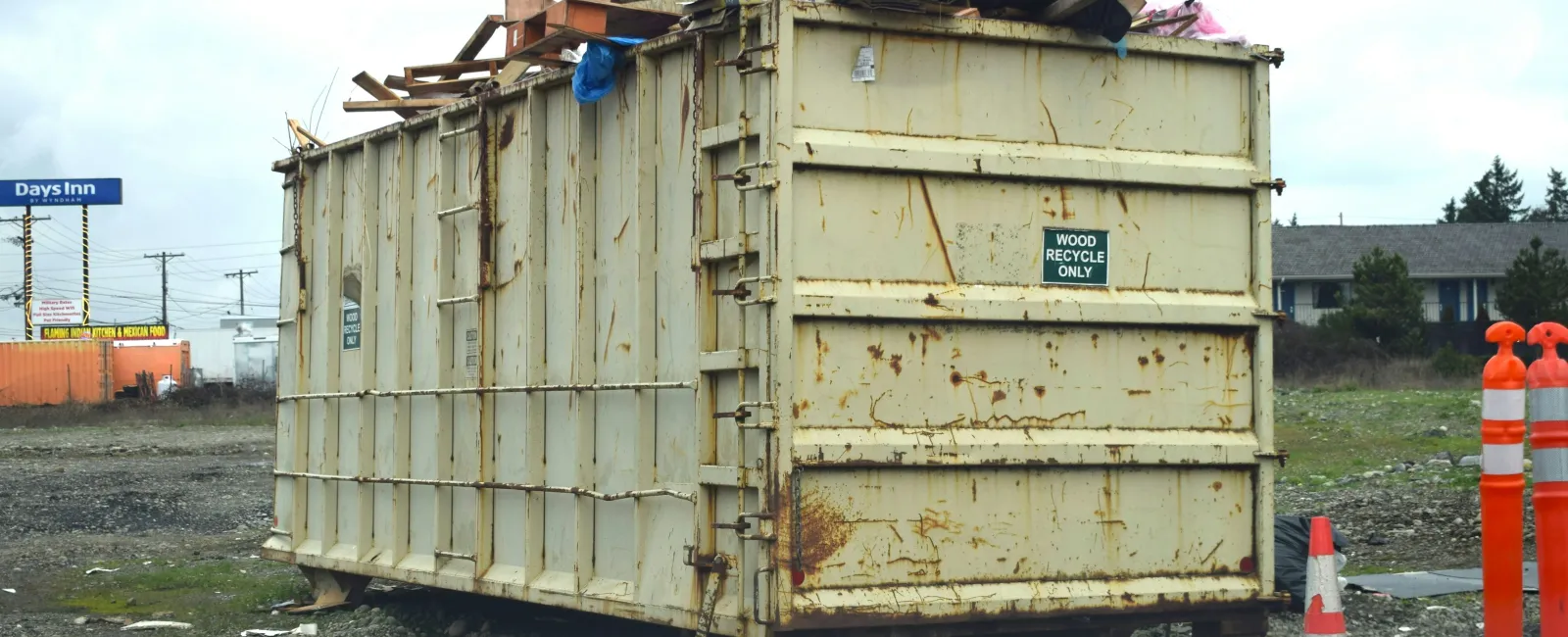Large rusty metal dumpster filled with wood debris, labeled for wood recycling, on gravel near traffic cones.