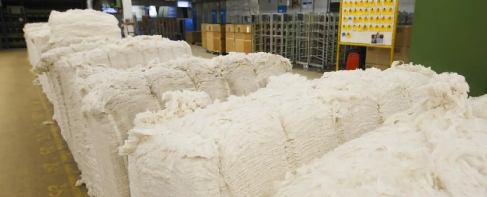 Large stacks of raw white cotton bales stored inside a warehouse with industrial shelving and crates.
