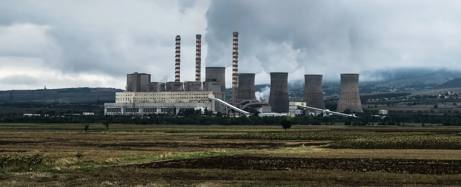 Industrial power plant with tall smokestacks emitting smoke under a cloudy sky near open fields.
