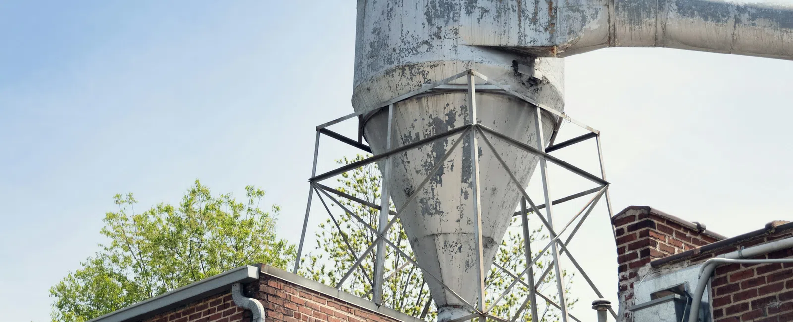 Rusty industrial dust collector mounted on brick building under clear sky with trees in background