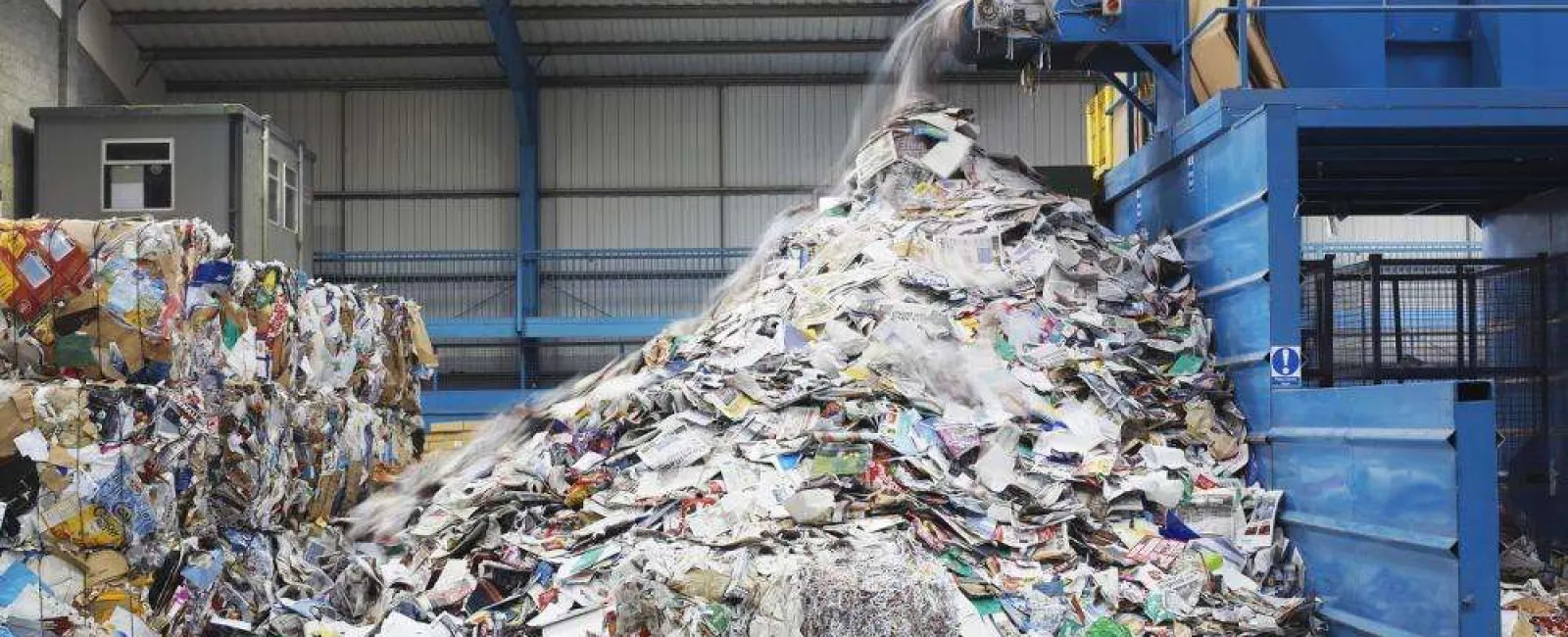 Industrial recycling facility with piles of sorted paper waste and large blue machinery processing materials