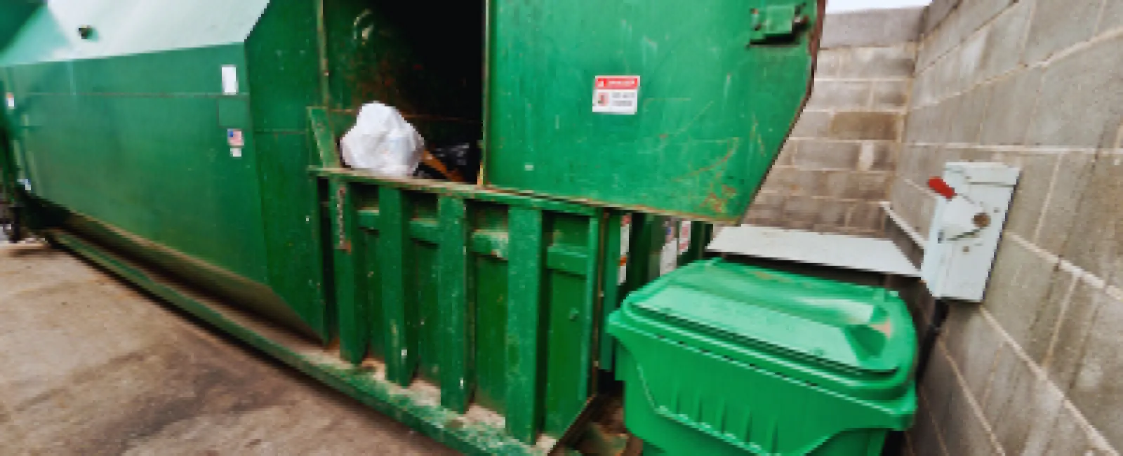 Large green industrial dumpster with lid open and a green waste bin beside a concrete block wall under metal beams.