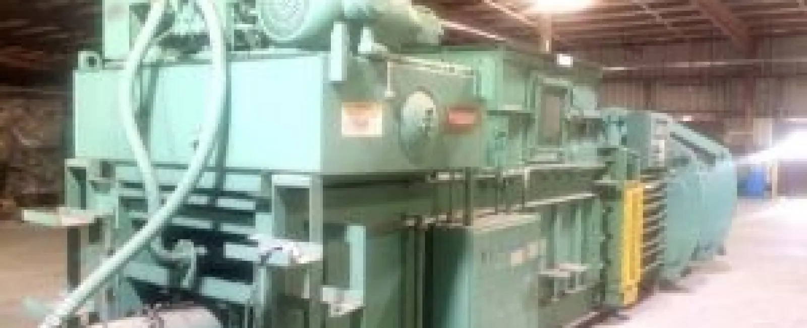 Large green industrial machine with pipes and motor inside a spacious warehouse with high ceiling and lighting.