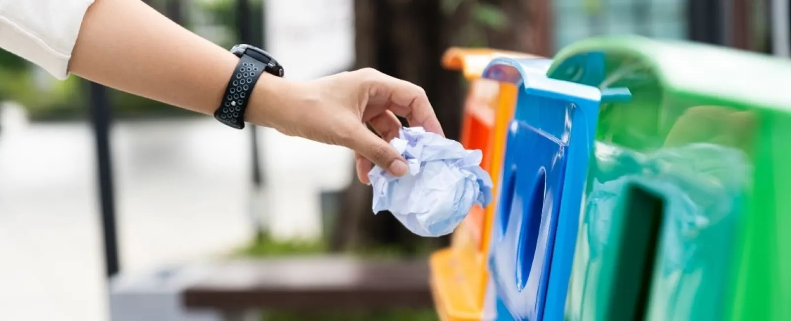 Person's hand throwing crumpled paper into a blue recycling bin outdoors with colorful bins nearby.