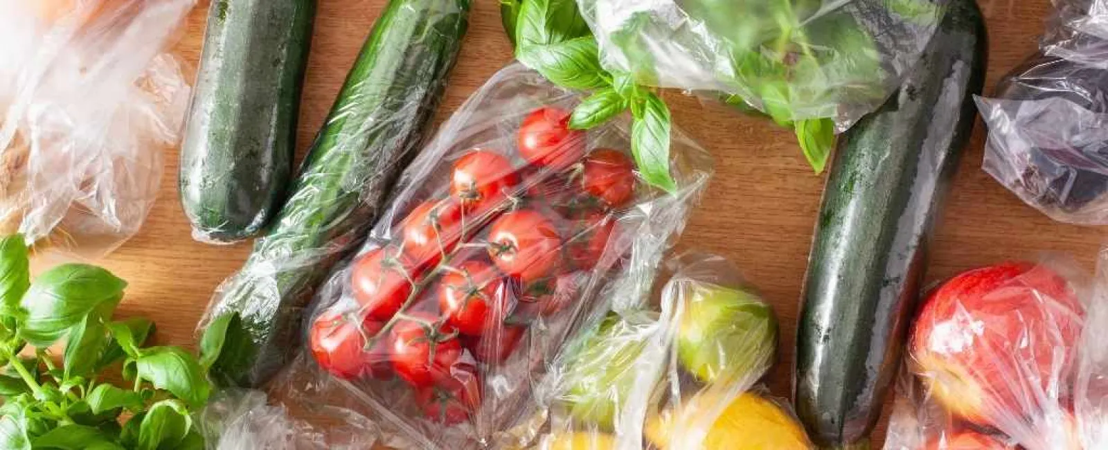 Fresh vegetables including zucchinis, cherry tomatoes, lemons, and herbs wrapped in clear plastic on a wooden surface