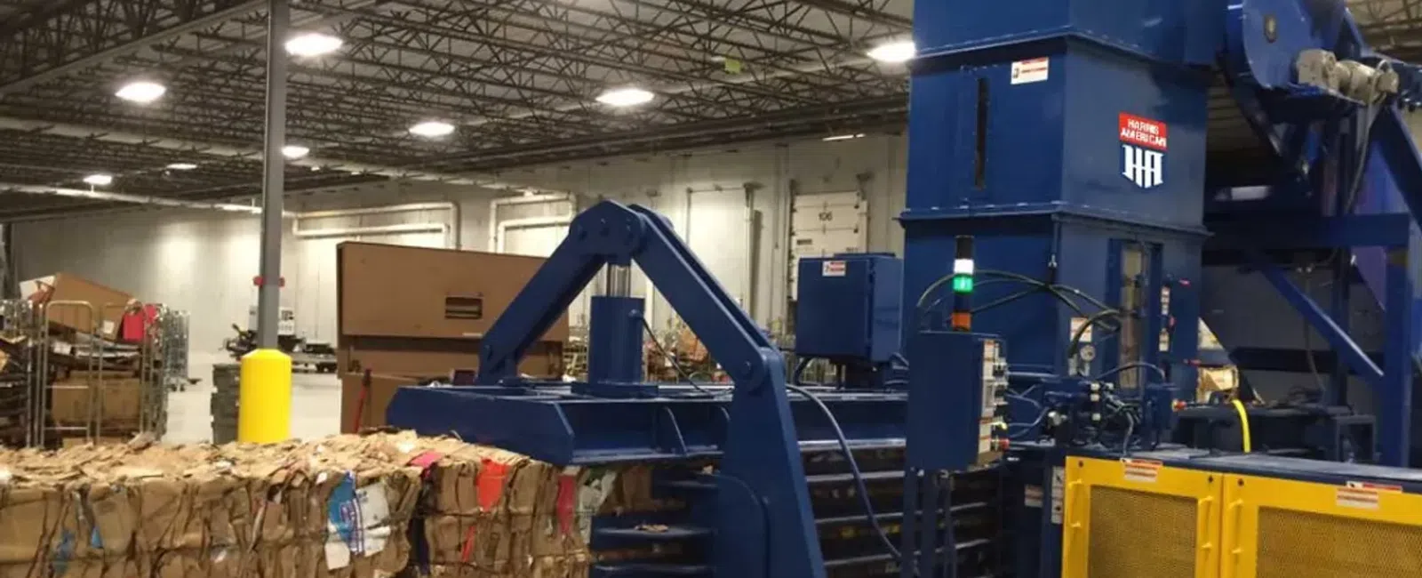 Industrial baler compressing cardboard in a large warehouse with stacks of recycled paper bales