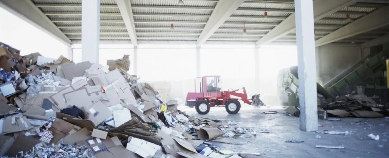 Interior of a recycling facility with piles of paper waste and a red loader vehicle in the background under a metal roof.