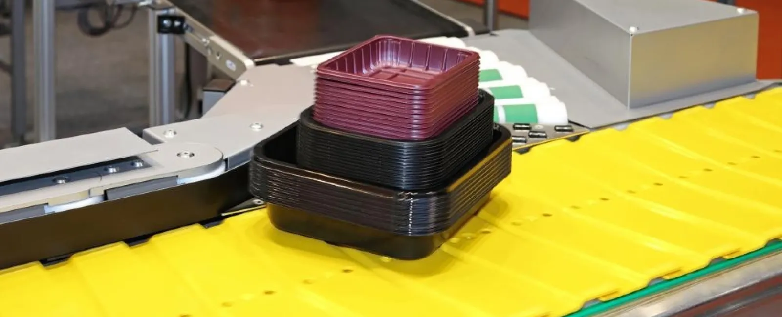Plastic trays stacked on a conveyor belt in a factory setting with yellow surface and machinery.
