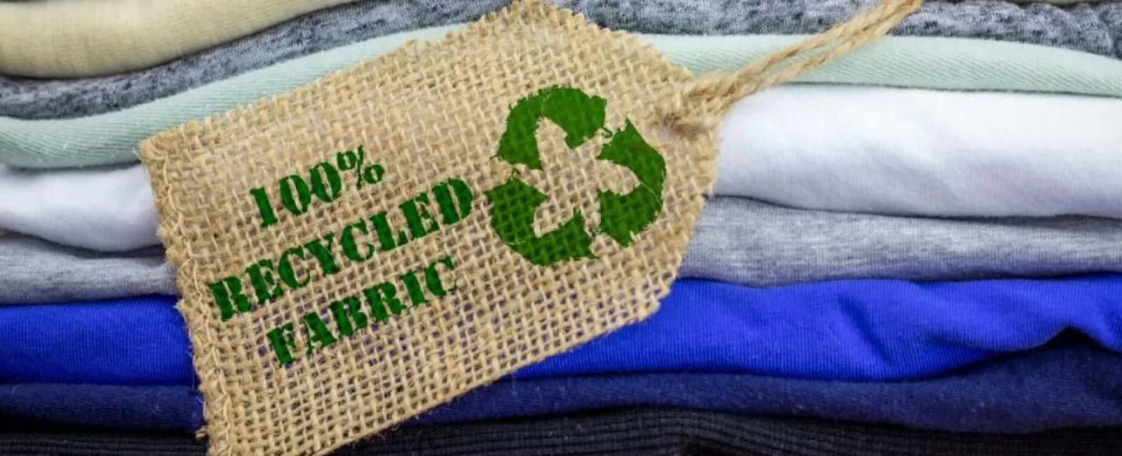 Stack of colorful folded clothes with a 100% recycled fabric tag featuring a green recycling symbol.