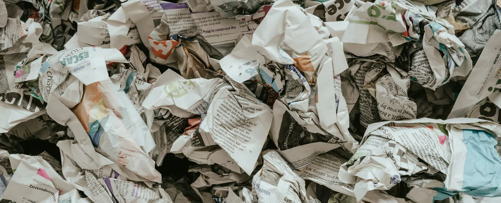 Pile of crumpled newspapers and printed paper waste in various colors and text scattered together.