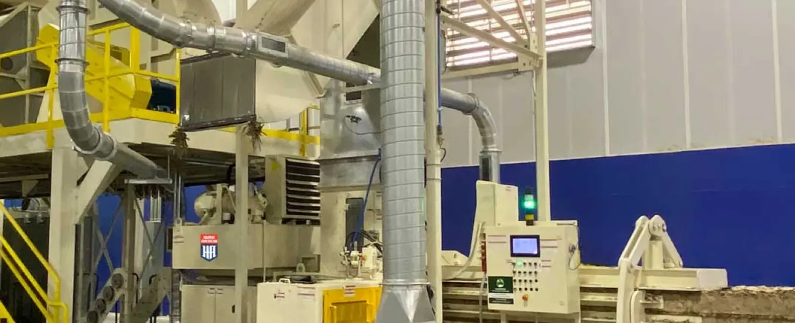 Industrial machinery in a factory with yellow stairs, metal ducts, control panel, and compressed material bales.