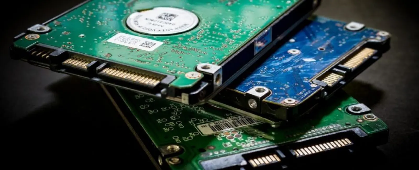 Close-up of stacked green and blue hard drives showing circuit boards and connectors on dark surface.