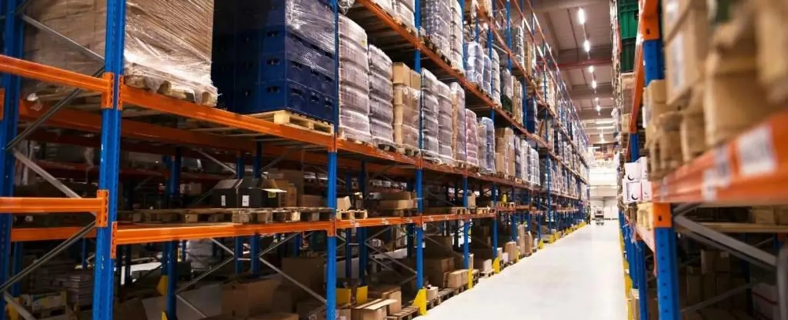 Warehouse aisle with tall shelves stocked with pallets of goods and boxes under bright lighting.