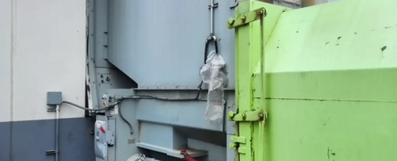Industrial waste compactor with large green container and mechanical components in facility corner.
