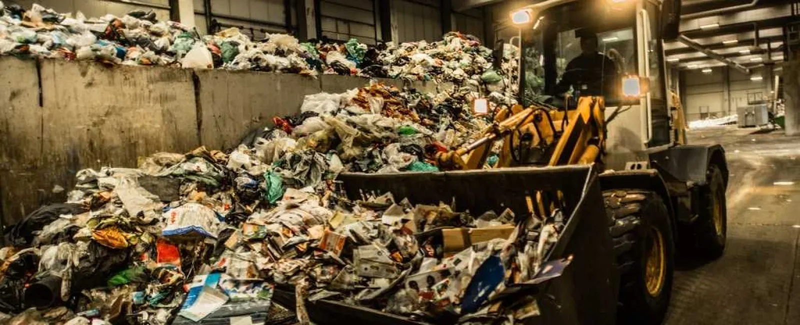 Heavy machinery loading large piles of mixed waste inside a dimly lit waste processing facility.
