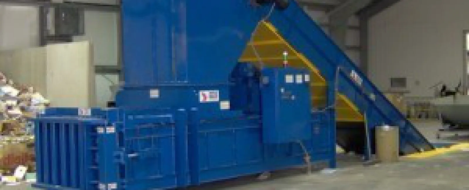 Large blue industrial baler machine with conveyor belt inside a warehouse for compacting materials.