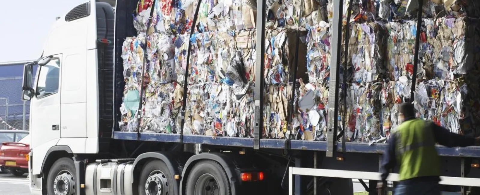 Truck loaded with large compacted bundles of recyclable paper and cardboard at a recycling facility with worker nearby.