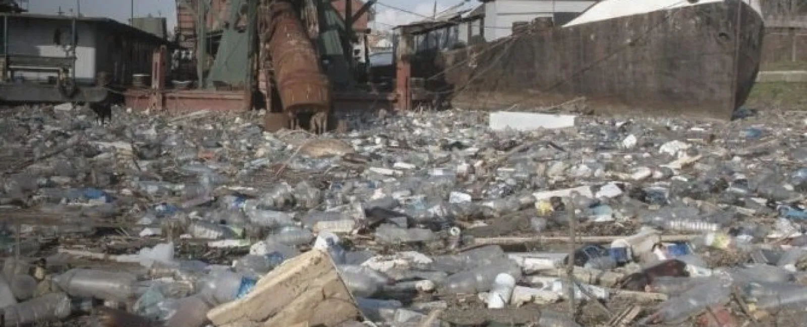 Large pile of plastic bottles and waste near industrial machinery and docked ships in polluted area.