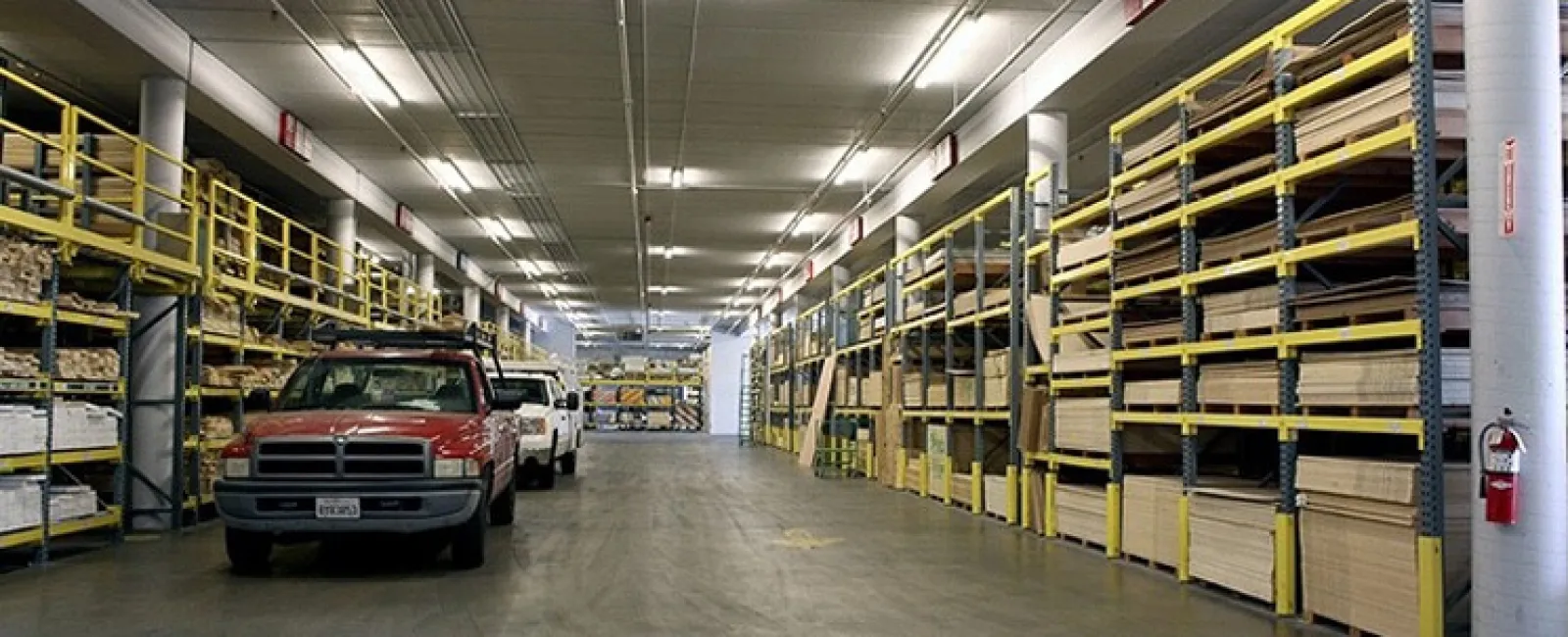 Interior of a large warehouse with yellow shelving stocked with wood panels and trucks parked inside.