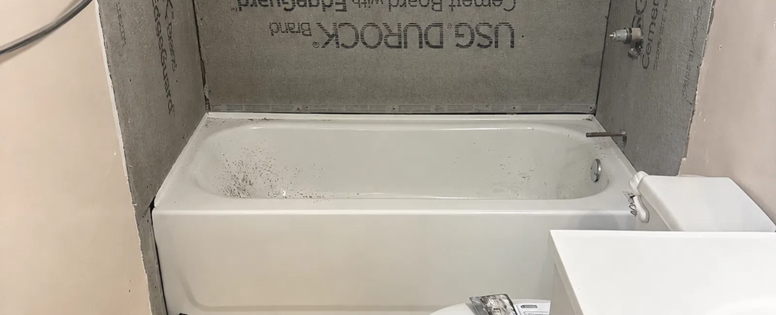 Small bathroom under renovation with cement board surrounding bathtub and tools on toilet seat.