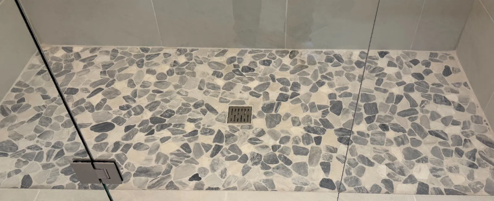 Modern shower area with pebble stone floor tiles and glass door enclosure with metal hinges and drain.