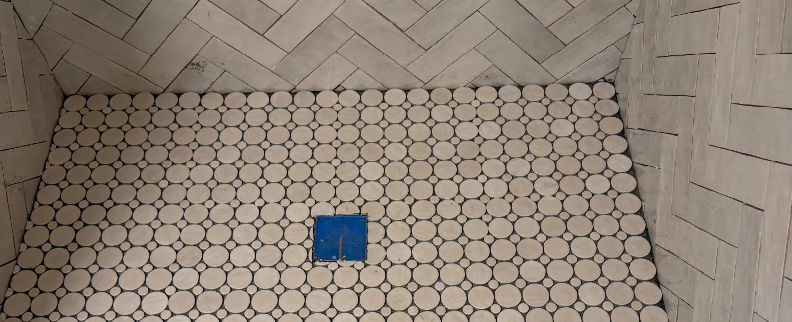 Shower floor with round white tiles and blue drain surrounded by beige herringbone patterned wall tiles.