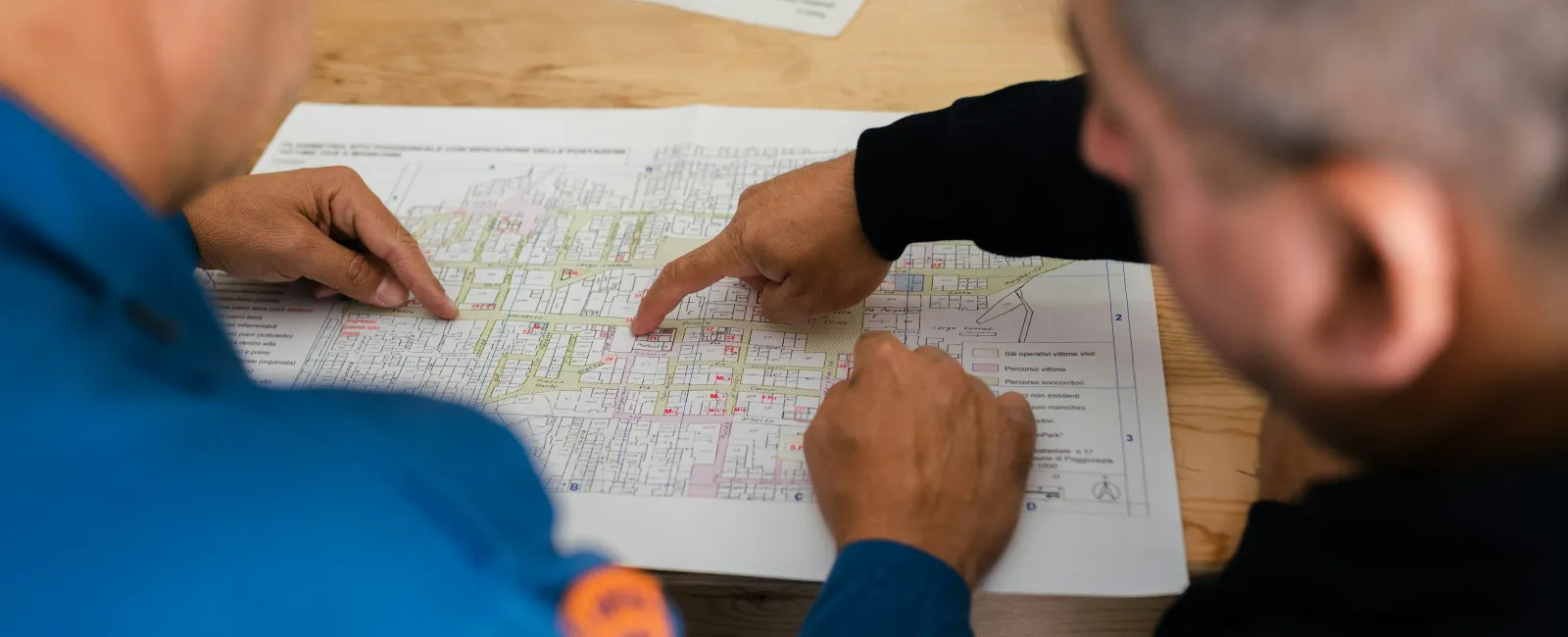 Two workers reviewing a detailed map or blueprint on a wooden table, pointing at specific areas.