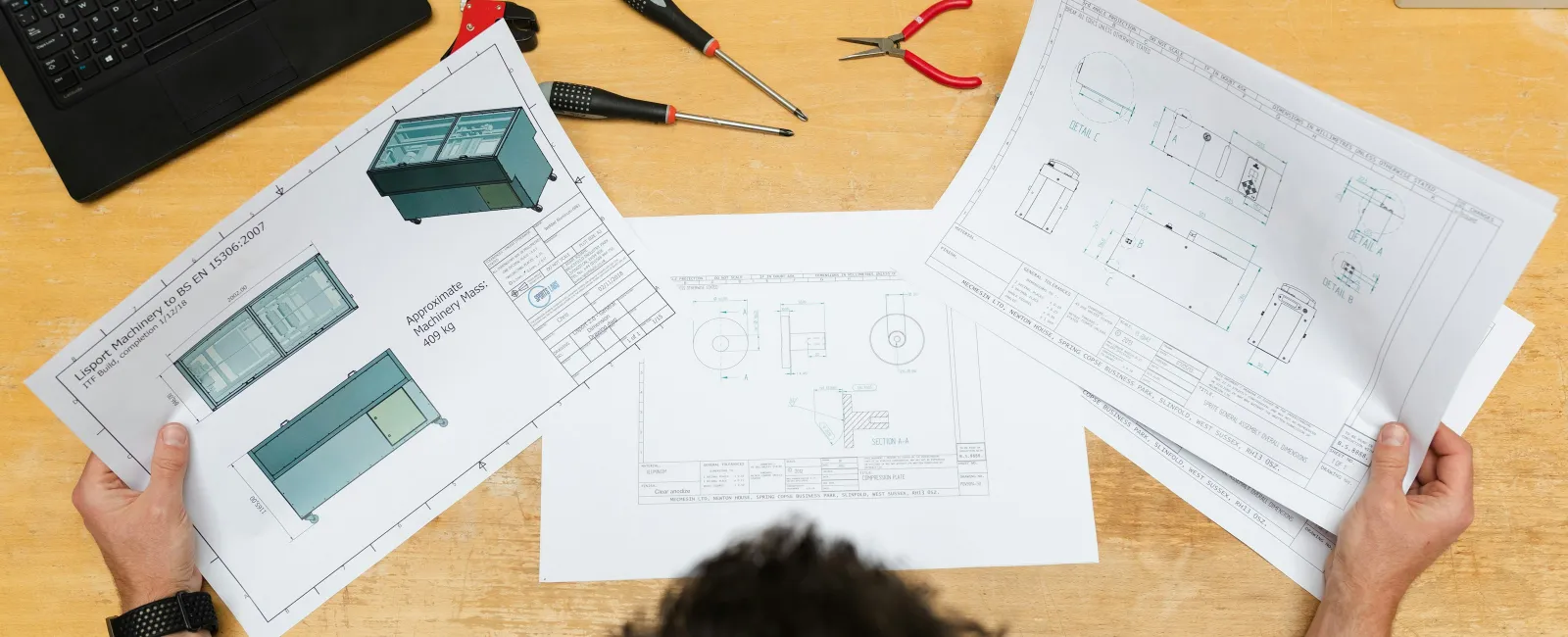 Man reviewing detailed technical blueprints and 3D design drawings at a wooden desk with tools and laptop