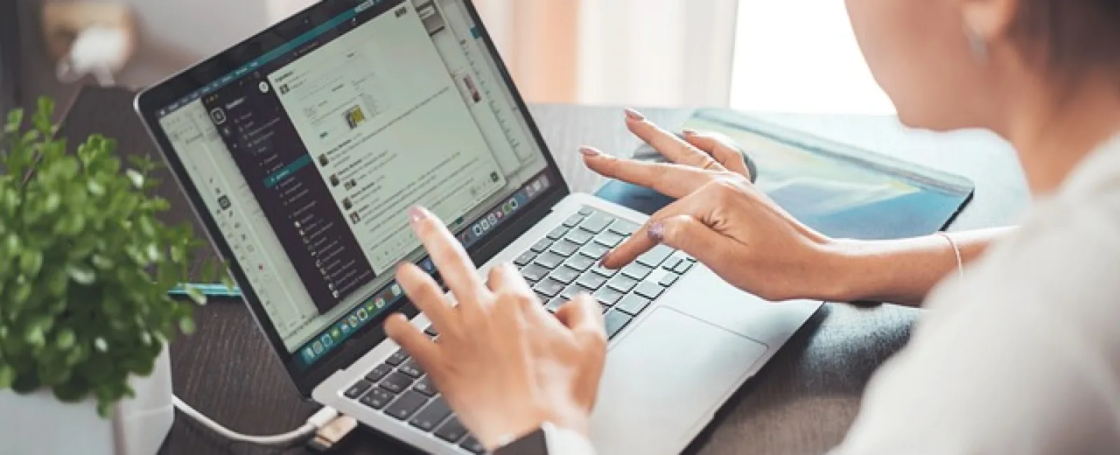 Woman using laptop messaging app at desk with smartphone and plant nearby in a bright room.