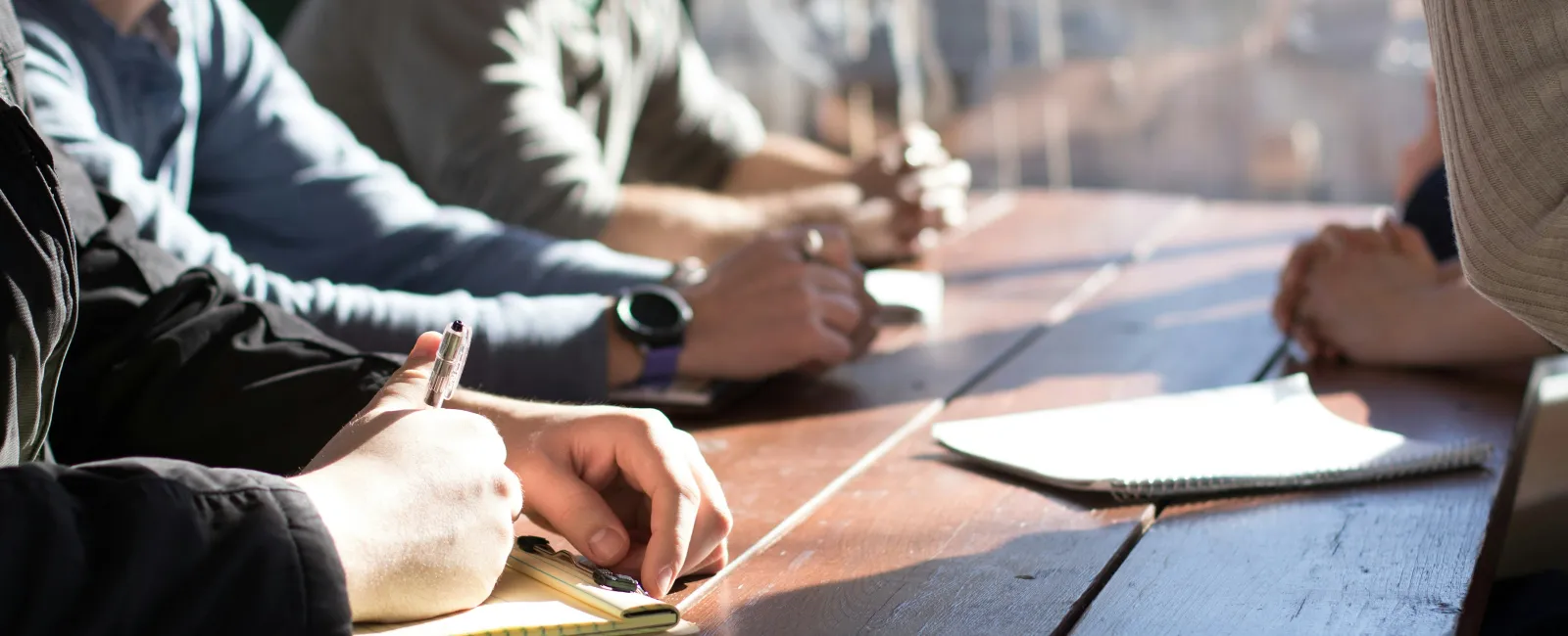 People sitting at a wooden table during a meeting, taking notes and discussing ideas in natural light.
