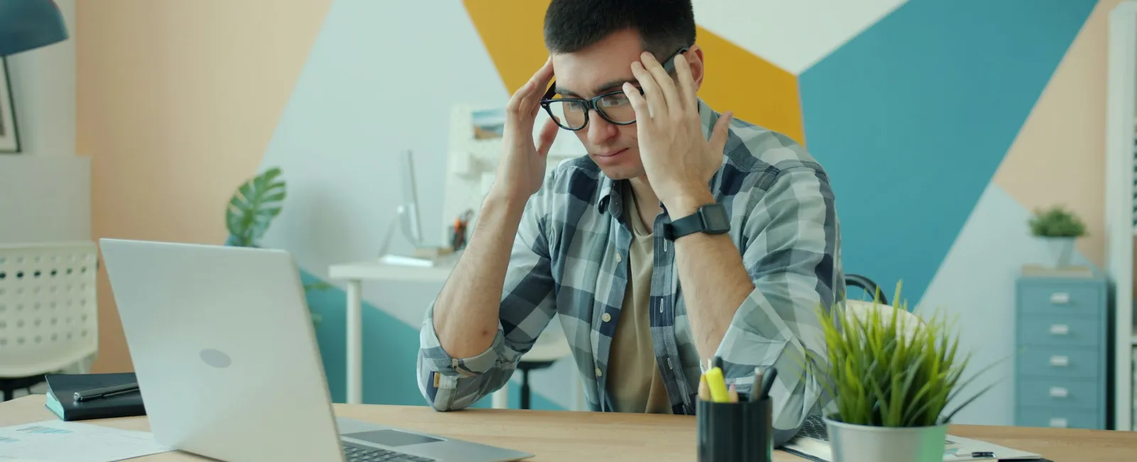 Young man wearing glasses stressed while working on laptop in a modern office with colorful geometric wall art.