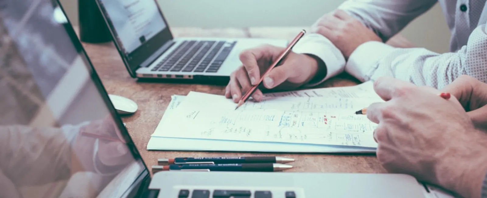 Two people working together at a desk with laptops and notes, discussing plans and ideas with pens and documents.