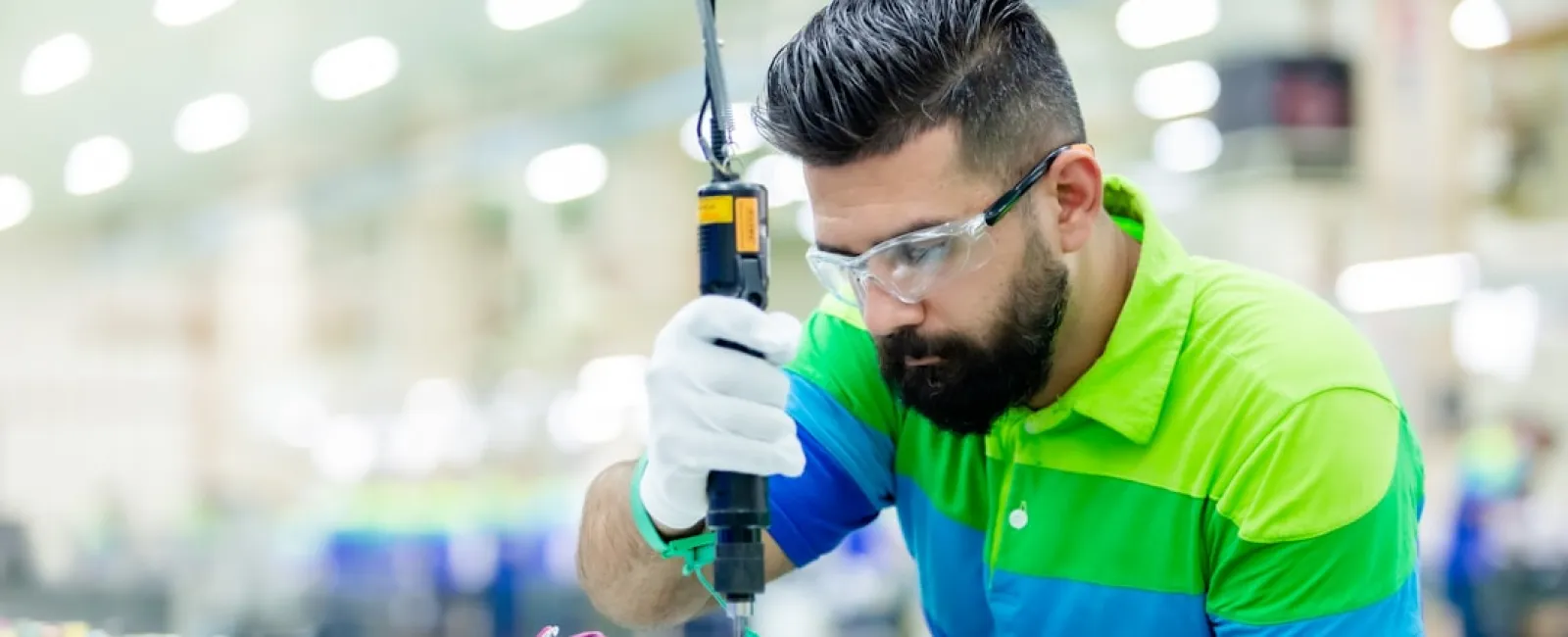 Technician wearing safety glasses and gloves assembling electronic circuit board in factory setting.