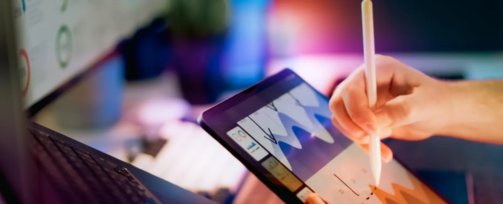 Person using stylus on tablet to analyze colorful data charts next to laptop in a modern work setup.