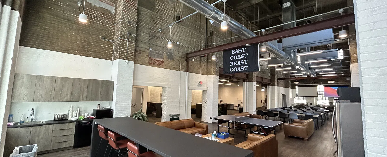 Spacious modern office lounge with high ceilings, industrial ductwork, kitchen area, bar seating, and ping pong tables.