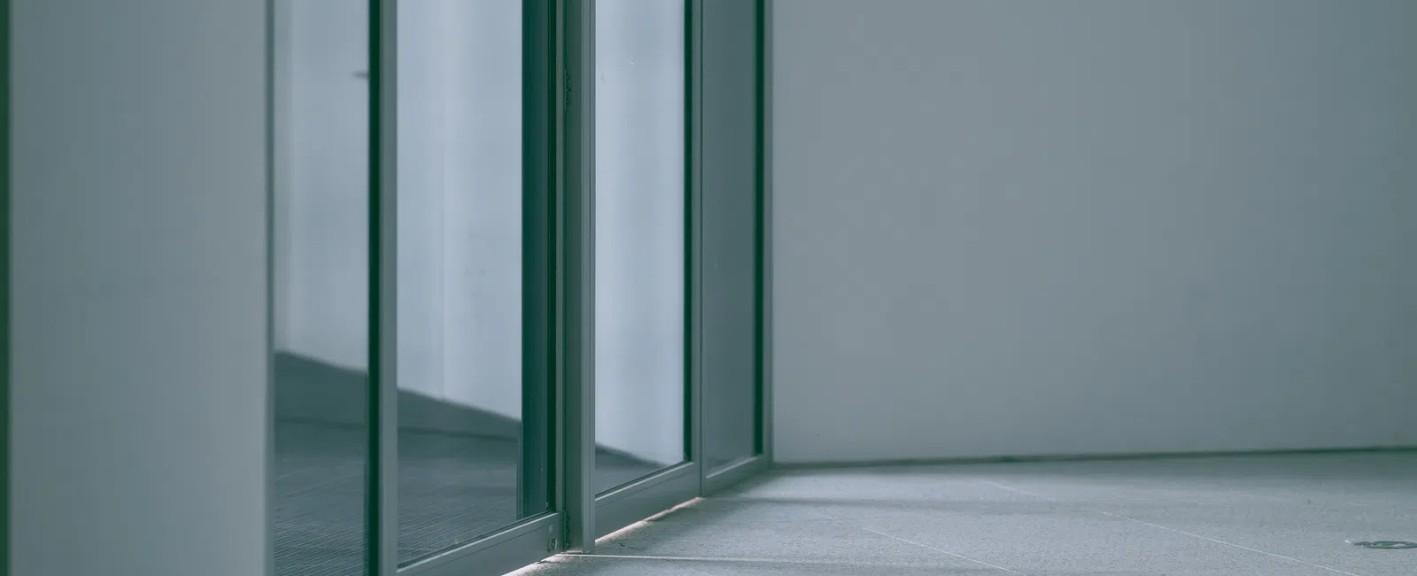Minimalist corner with glass panels reflecting light on a smooth tiled floor in a modern building.