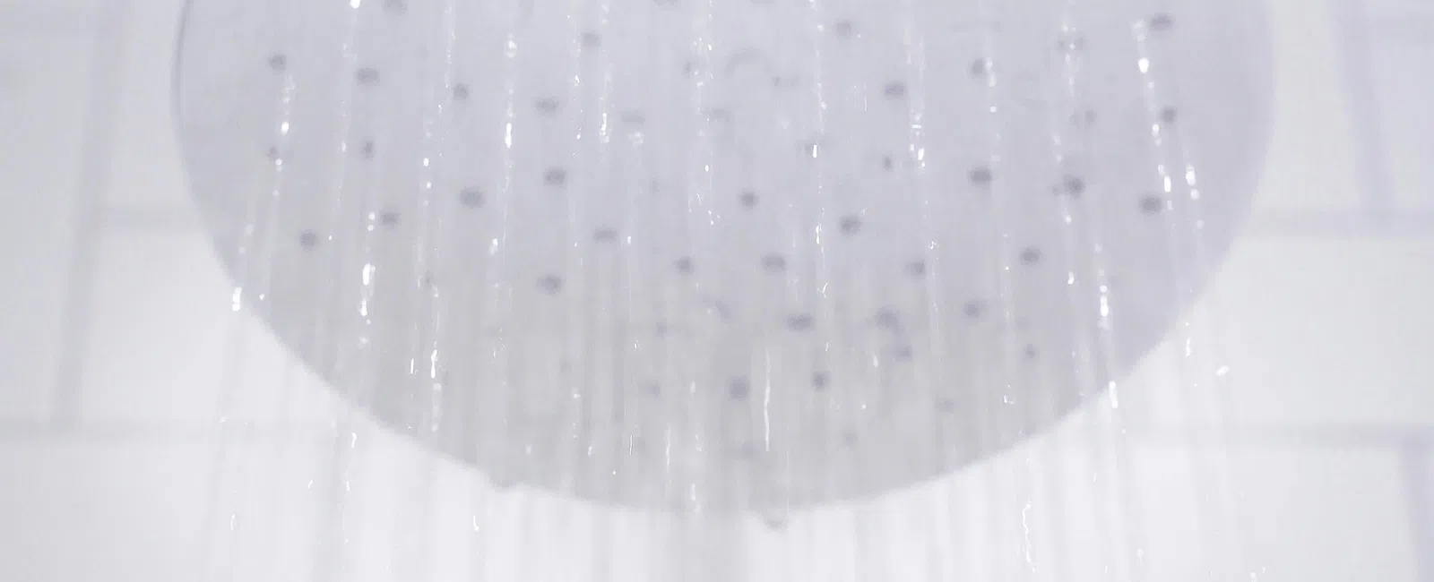 Round metal showerhead with water flowing down against a white tiled bathroom wall.