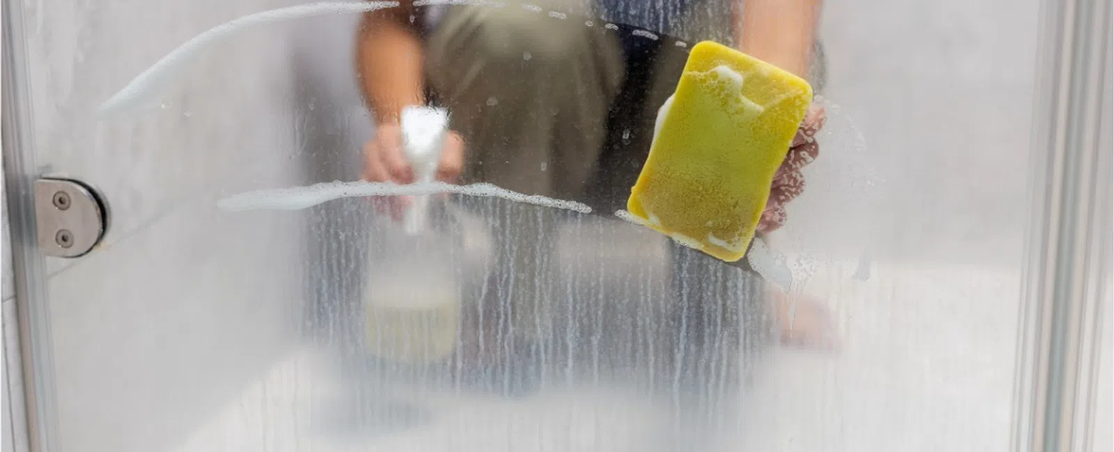 Person cleaning a foggy glass shower door with a yellow sponge and spray bottle in bathroom.