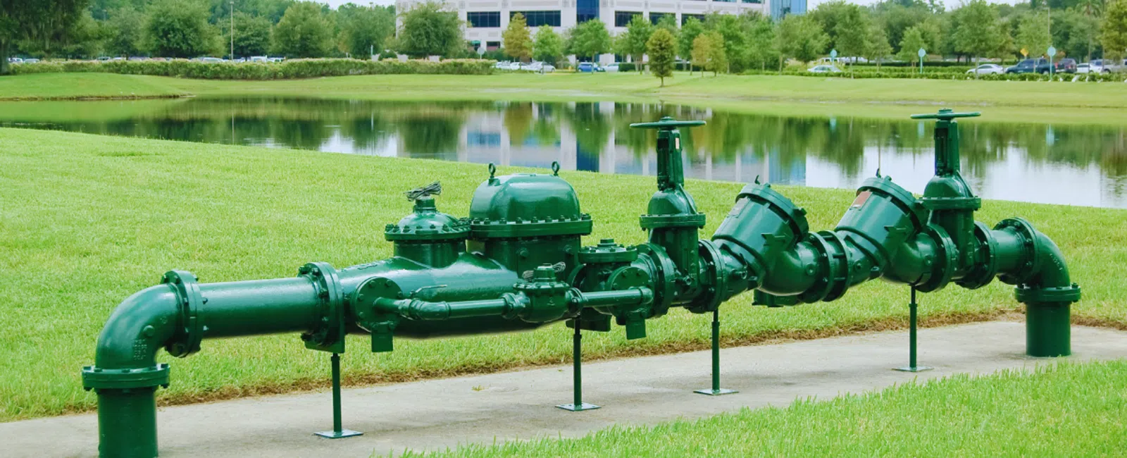 Green industrial water pipeline with valves on grassy lawn near lake and office building in background
