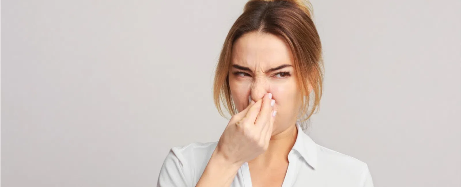 Woman in white shirt holding nose with a disgusted expression against a neutral background.