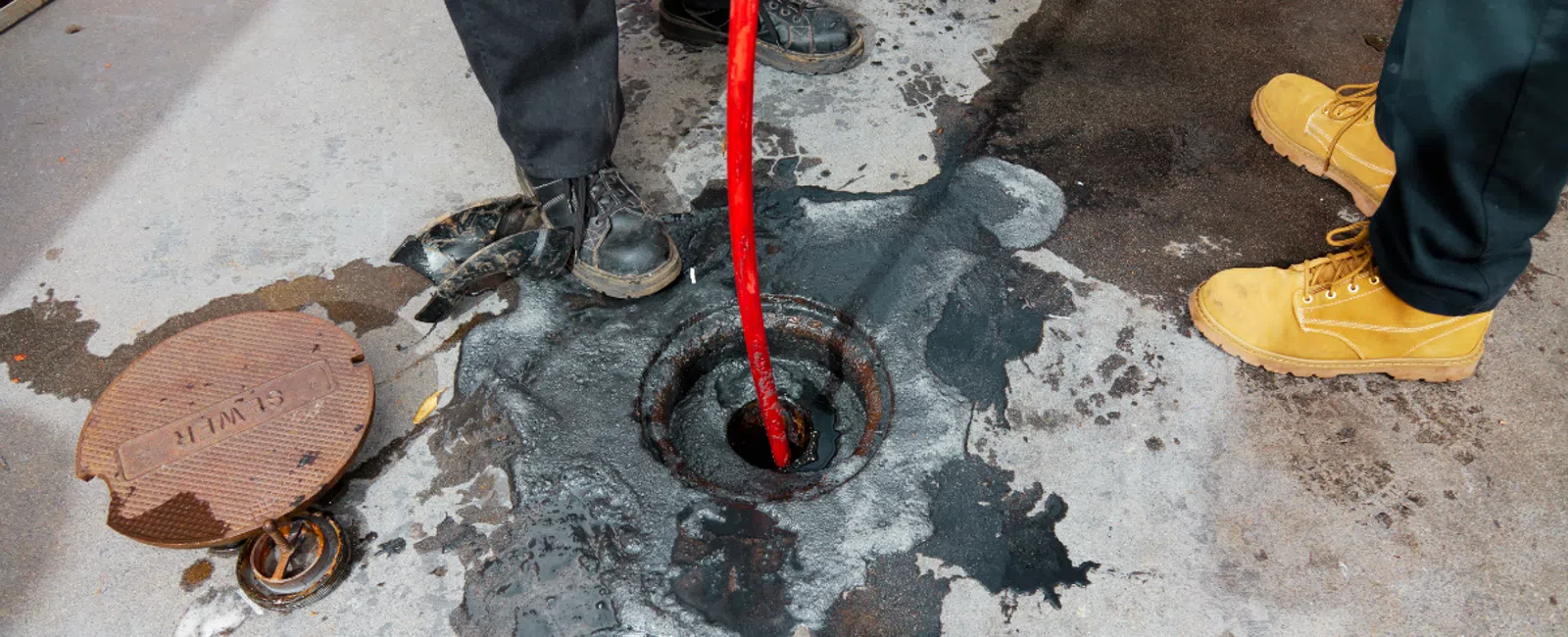 Workers removing blockage from a manhole with a red hose on a worn concrete surface.