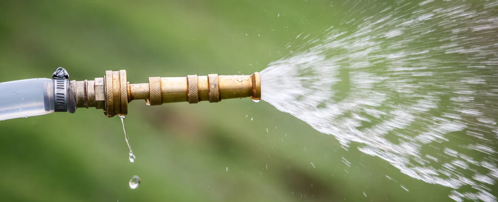 Close-up of a garden hose nozzle spraying water with droplets falling against a blurred green background.