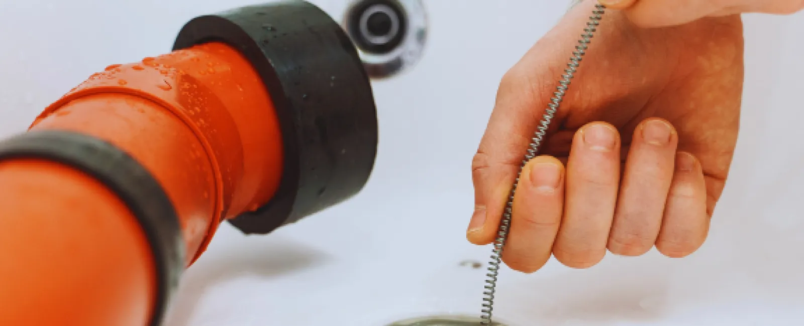 Hand using a drain snake tool to unclog a bathtub drain next to an orange pipe fitting.